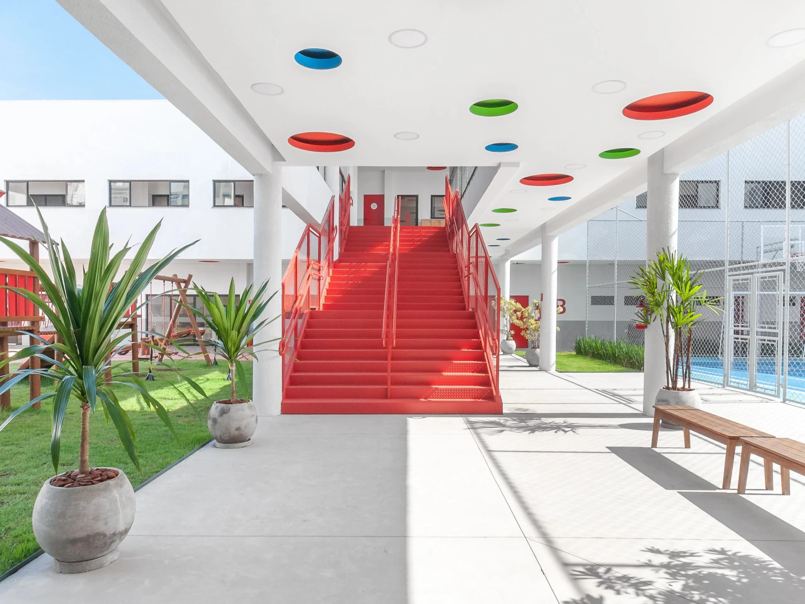 Ambiente escolar com arquitetura escolar planejada, onde apresenta um pátio de escola com uma escada vermelha e detalhes no teto, com circulos coloridos.
